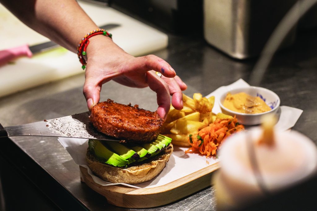 A chef plates up a vegan patty onto a burger bun with avocado and a side of french fries at a restaurant. 
