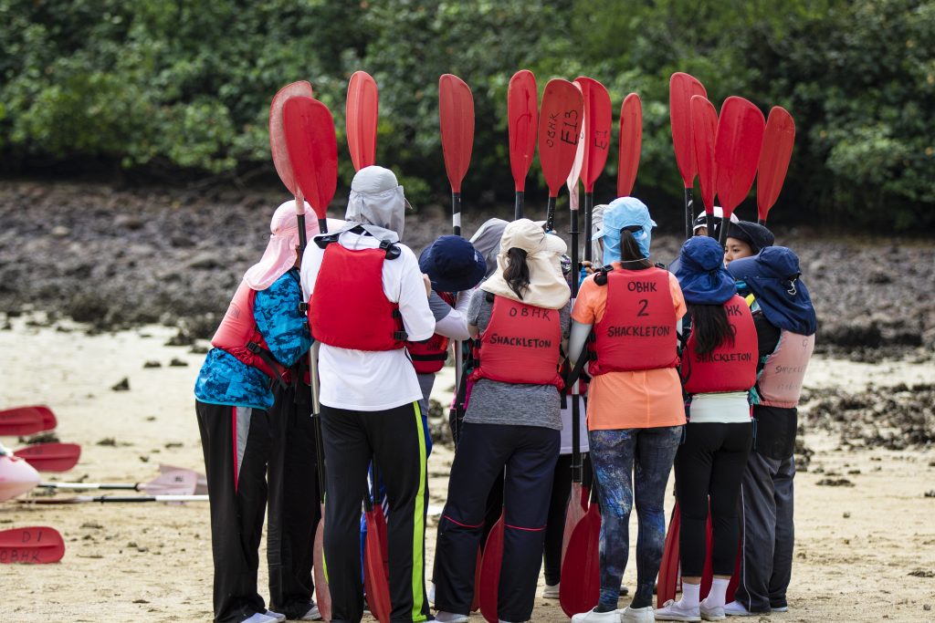 A group of young people stand on a beach with paddles in their hands in a group as part of a teamwork exercise.