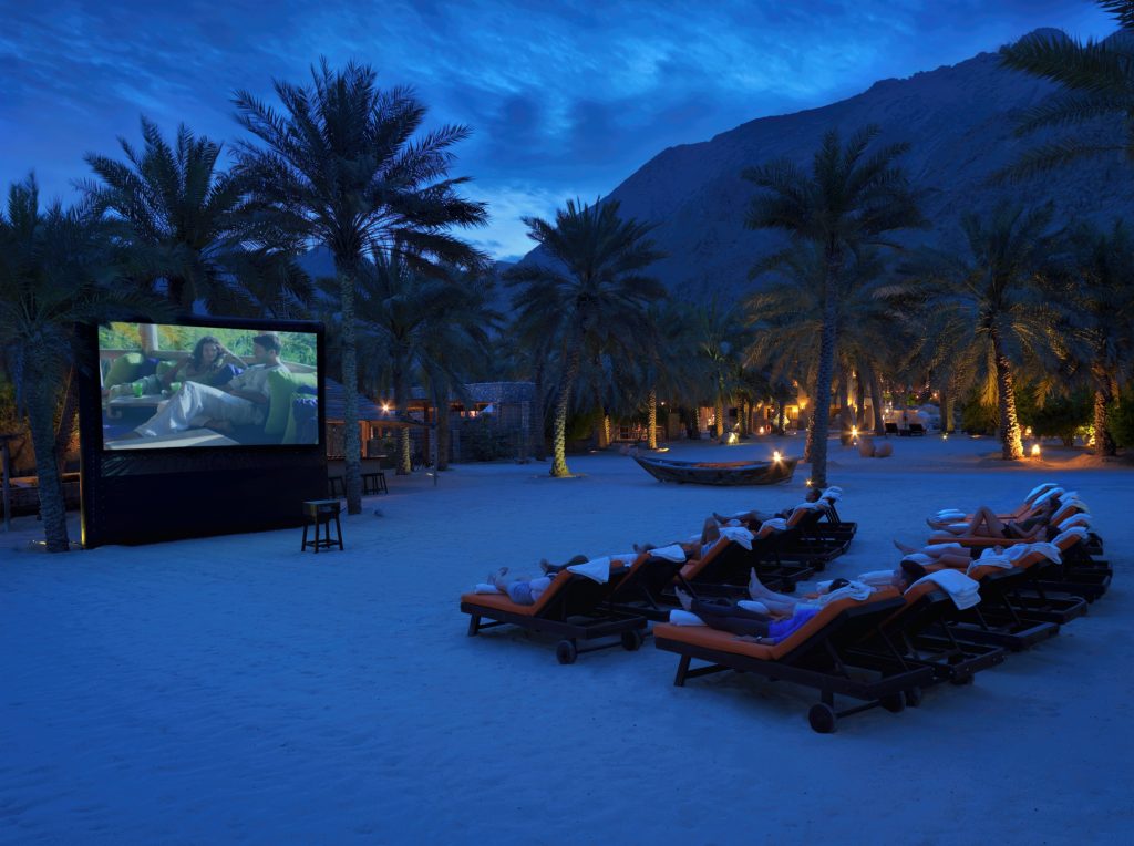 Several people watch a large outdoor television screen showing a film at the outdoor cinema at Six Senses Zighy Bay in Oman.