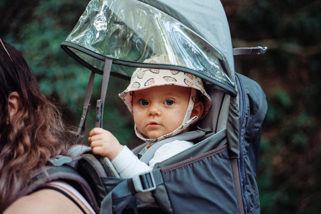 A  baby sits inside a baby carrier for hiking, covered with a rain cover. 